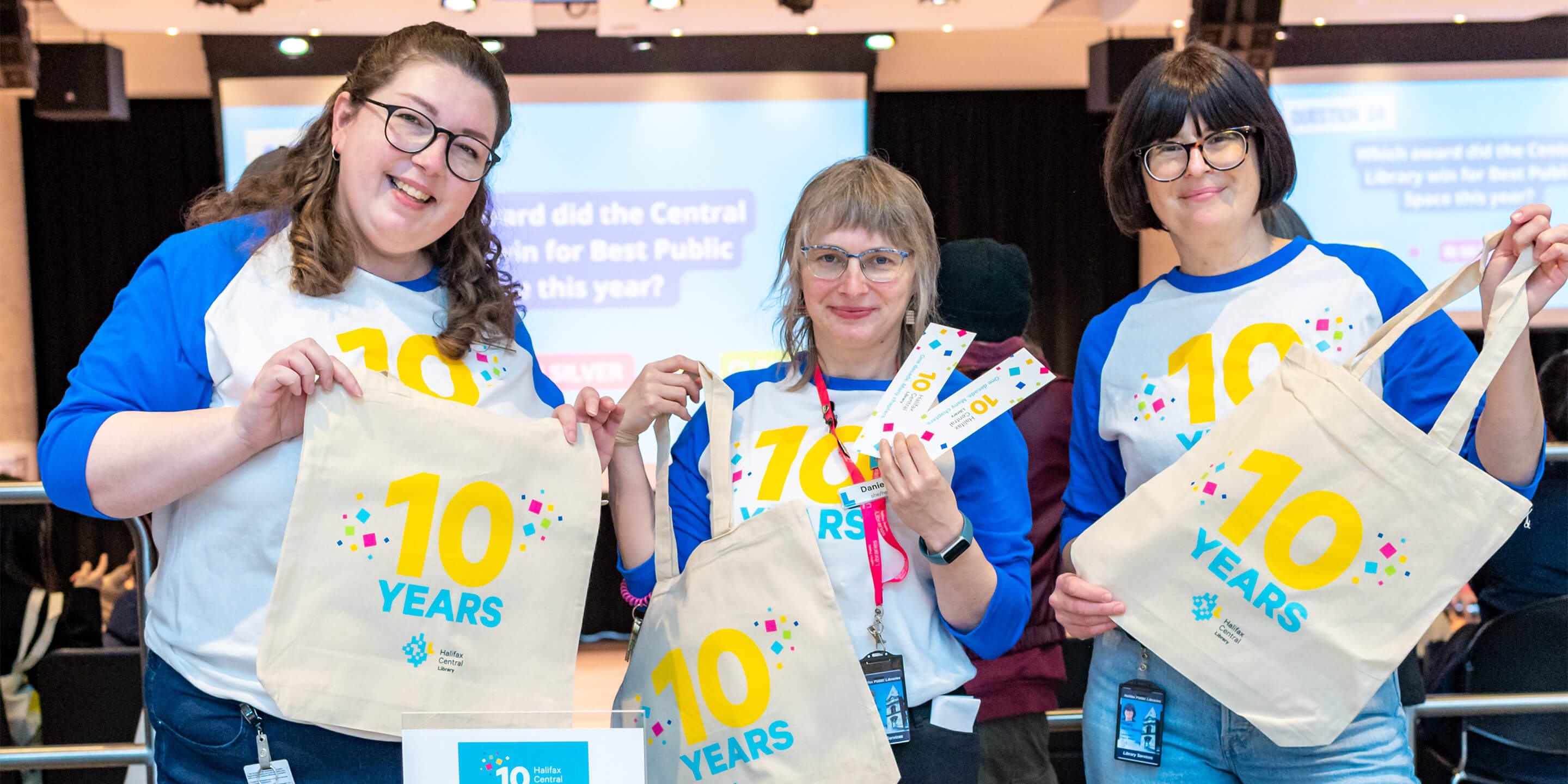 Staff holding 10 year anniversary tote bags while wearing matching anniversary shirts, smiling to camera.